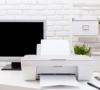 A white printer next to a computer in front of a white brick wall