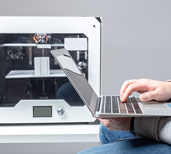 a person on their laptop that is connected to a futuristic 3D printer.
