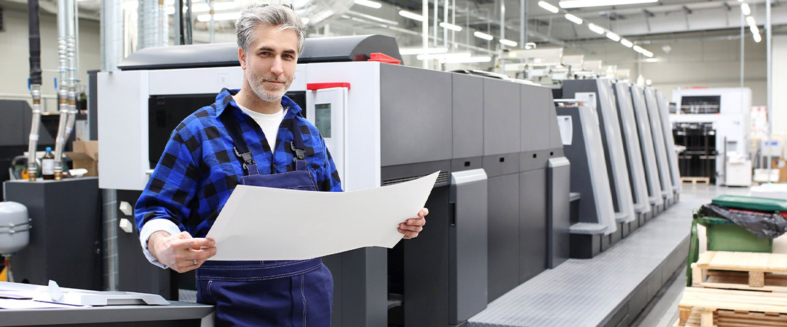  A person in overalls holding a blueprint in front of an industrial printer