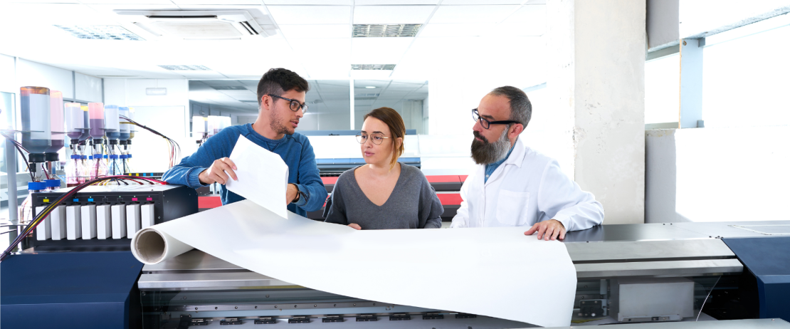 Three professionals stand over a large format printer and a large blank sheet of paper